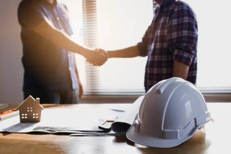Homeowner and construction professional shaking hands beside plans and a hard hat